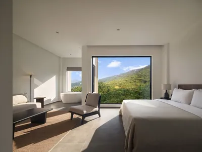 簡約臥室內設有大型窗戶，窗外可見綠意盎然的山坡景觀，室內擺放木質床架搭配白色床品，以及陶瓷地磚。