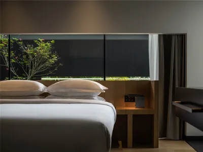 A minimalist bedroom with a wooden bed, white bedding, and a glass window with a tree outside.