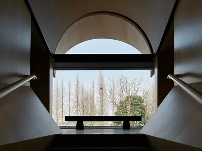 Concrete staircase viewed from above, featuring an arched window with a bench and a scenic outdoor view of bare trees.