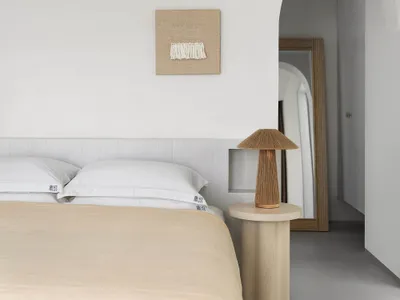 A minimalist bedroom with neutral-toned bedding and a wooden side table with a lamp.