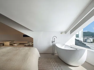 Modern minimalist bedroom with white concrete walls and floor, large window with city view.