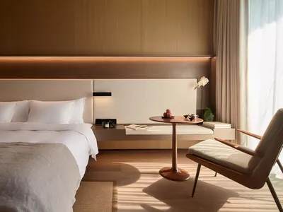 A minimalist bedroom featuring a bed with wooden headboard, white bedding, and a wooden side table with a book and coffee cup