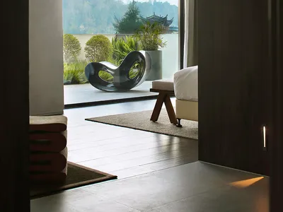 A modern minimalist bedroom with wood flooring, a white bed, and a black sculptural chair on a balcony overlooking a scenic l