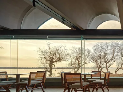 Interior view of a modern restaurant featuring large glass windows, concrete columns, and wooden tables and chairs.