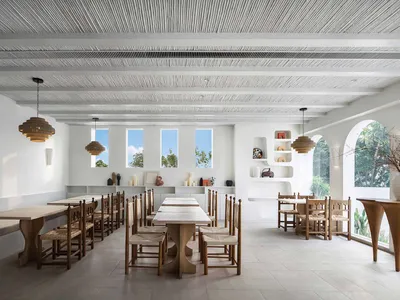 A minimalist restaurant interior featuring white concrete walls, wooden tables and chairs, and concrete floors.
