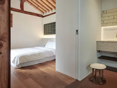 Minimalist bedroom with white walls, light wooden floor, white bedding, and a wooden stool under a granite countertop with a