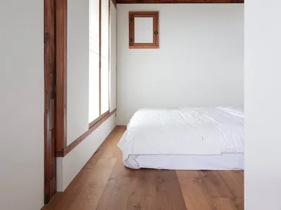 A minimalist bedroom with white walls, wooden floors, and a single white bed against a white wall. Wooden beams in the ceilin