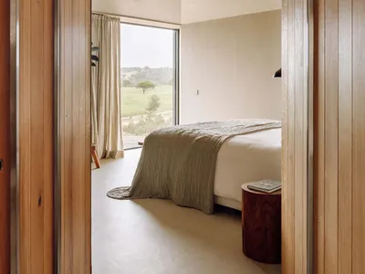 A minimalist bedroom featuring a bed with a wooden frame and a concrete floor, with a view of a natural landscape through a l