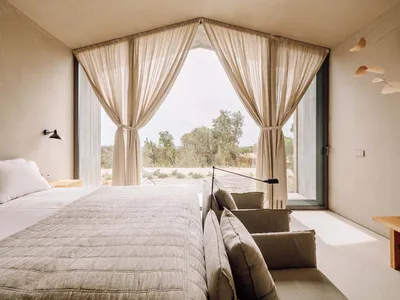 A minimalist bedroom with fabric curtains and a concrete floor, featuring a comfortable bed and a small seating area.