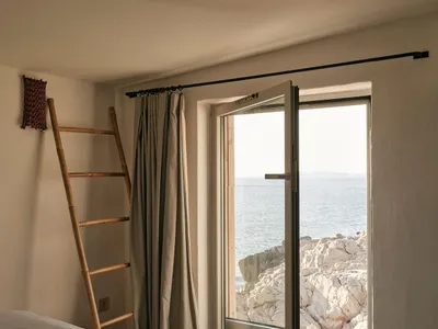 A minimalist bedroom featuring a stone construction, with a window overlooking the sea and rocks, and a bed with a white shee