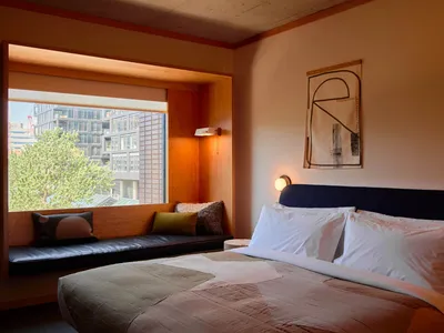A minimalist bedroom featuring a bed with white linens, wood furniture, and a window seat with pillows.