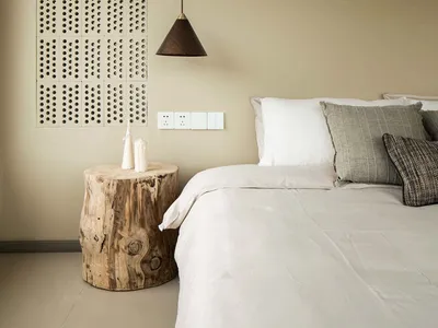A minimalist bedroom featuring a wooden stump nightstand with a single candle holder and a modern pendant light hanging above