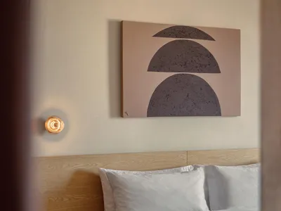 A minimalist bedroom with a wooden headboard and white bedding, featuring a wall-mounted light fixture and a large abstract p