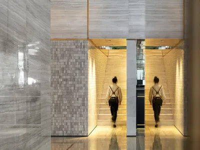 Two people walking up the stairs in a modern hotel lobby with marble walls and glass railings.