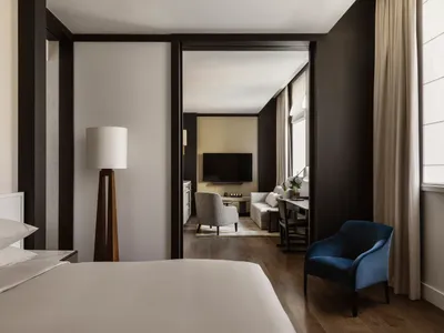 A modern hotel room featuring a minimalist bedroom with a bed, lamp, and a door leading to a living space.