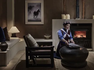 A cozy living room with a fireplace, wooden beams, and a woman reading by the fire.