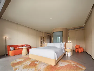 A modern minimalist bedroom featuring a bed with a wooden frame, white bedding, and an orange couch with pillows under a beig