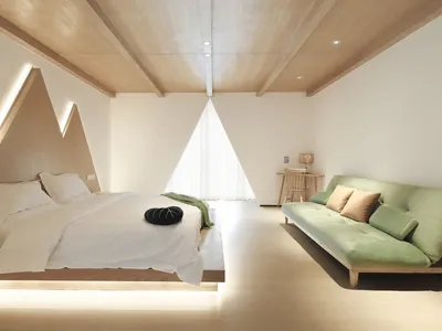 A minimalist bedroom featuring a bed with a geometric headboard and a green couch, set against a white wall and a wooden ceil