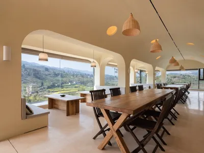 A spacious modern restaurant interior with wooden tables and chairs, large arched windows, and a view of the valley.