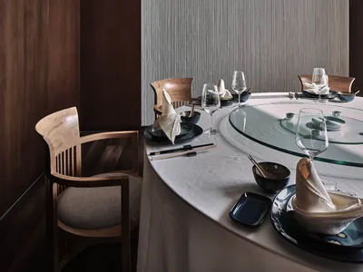 A minimalist restaurant dining room with dark wood walls, glass table, textured fabric chairs, and a woven rattan basket pend