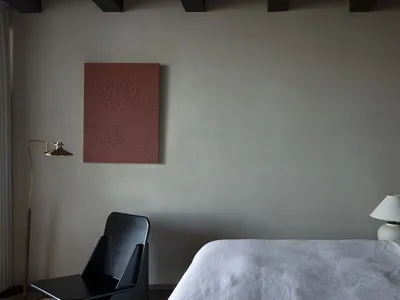 A minimalist bedroom featuring a white sheet on a bed with a black chair, a lamp, and a framed abstract painting on the wall.