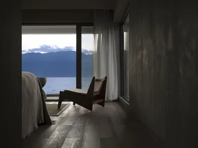 A minimalist bedroom with a view of mountains and water through large windows, featuring a wooden chair and natural lighting.