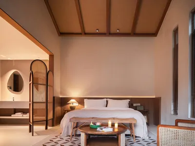 A minimalist bedroom featuring a large bed with a wooden headboard, a wooden bedside table with a lamp, and a wooden armchair