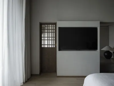 A minimalist bedroom with a wooden door featuring a geometric pattern and a flat-screen TV mounted on a white wall.