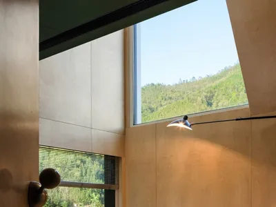 Simple, minimalist bedroom featuring a wooden platform bed with a green blanket and a book on top, large windows with a view