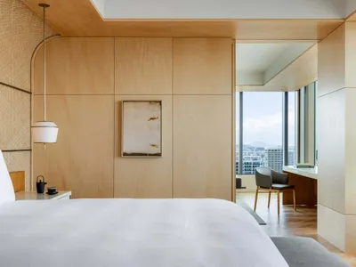 A modern bedroom with a bed, a chair, and a desk overlooking a cityscape.