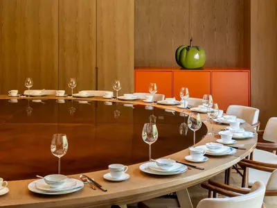 A modern restaurant interior with wooden furniture, featuring a large round table set for dining, a golden chandelier hanging