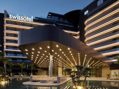 A modern hotel building with a curved glass facade illuminated at night, featuring a water feature and landscaping.