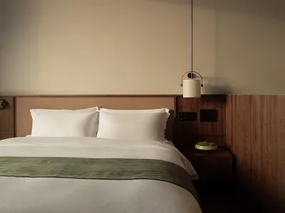 A minimalist bedroom with a wooden headboard and white bedding, featuring a hanging lamp.