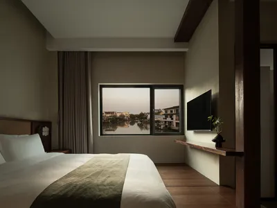 A minimalist bedroom featuring a comfortable bed and a window overlooking a serene waterway and residential buildings.