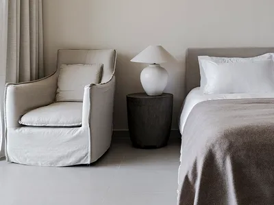 A minimalist bedroom with a bed, nightstand, lamp, and armchair in neutral tones.