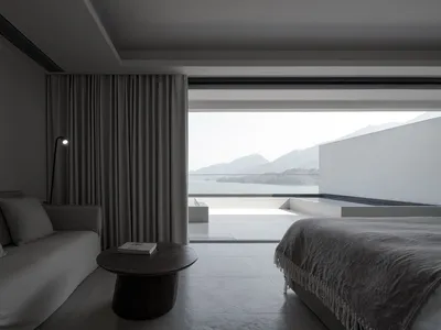 Modern minimalist bedroom featuring a large window with a view of mountains, a bed with a textured blanket, a couch, and a sm