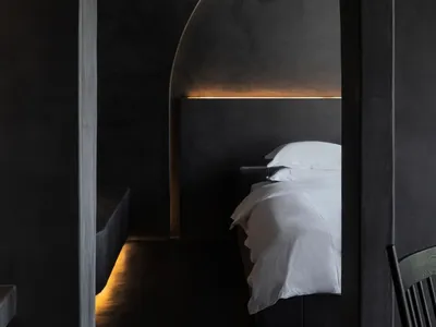 A minimalist bedroom with a concrete headboard and floor, featuring a single white bed and soft lighting.