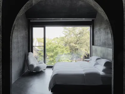 A minimalist bedroom featuring white bedding and a concrete interior, with a view of green trees outside through the window.