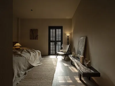 A minimalist bedroom with a bed covered in a textured blanket, a modern chair, and a wooden bench against a light-colored wal