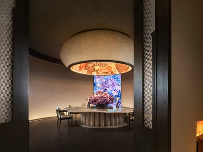 Modern restaurant interior with wooden furniture and a large round lamp hanging from the ceiling. Motion blinds are visible i