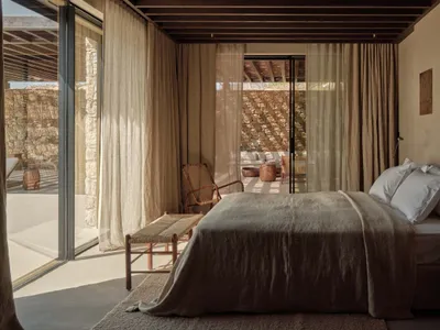 A minimalist bedroom with linen bedding, wooden furniture, and a stone wall, featuring a view blocked by curtains.