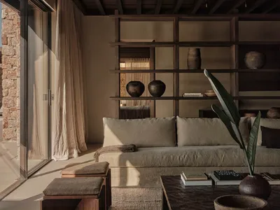 A modern, minimalist living room with wooden furniture, stone walls, and a large window allowing natural light to illuminate