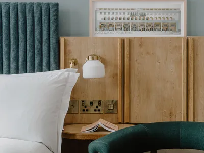 A minimalist hotel room featuring a bed with a wooden headboard and a wooden nightstand with a lamp, a chair with a wooden ar