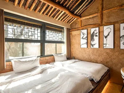 A bedroom with clay walls, wooden beams, and a large window with a view of the outdoors.
