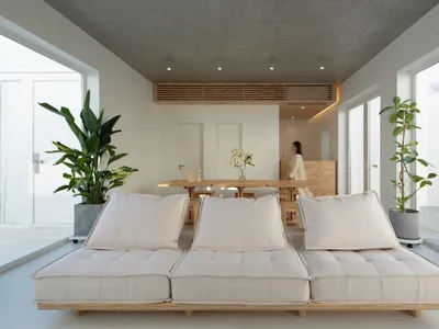 A minimalist interior space featuring a low-slung, cushioned sofa with light-colored cushions and a wooden dining table in th