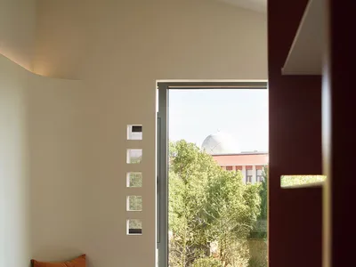 A minimalist bedroom featuring a concrete bench with a cushion and a window overlooking trees and a building.