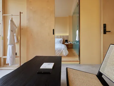 A minimalist bedroom with clay furnishings and light-colored walls, featuring a bed, desk, and chair.