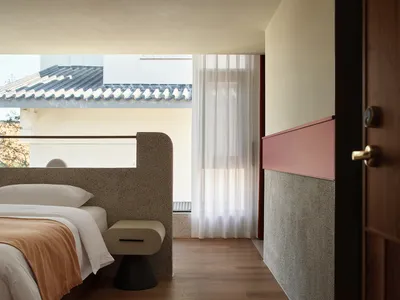 Minimalist bedroom with concrete and wood finishes, white bed, sheer curtains, and a view of a tiled roof.