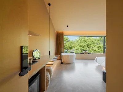 Modern minimalist hotel suite with concrete walls and floor, featuring a bathtub overlooking greenery.
