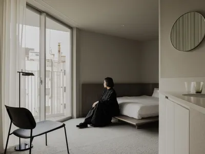 A minimalist bedroom with white furniture and a wooden bed, featuring a large window and a black chair.